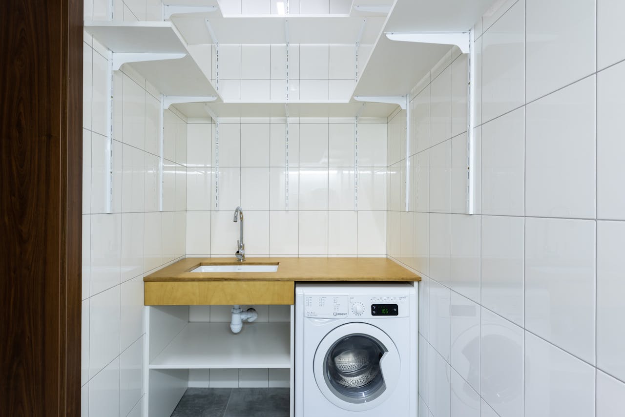 pexels-photo-7195881 Modern laundry area with white tiled walls, washing machine, and built-in shelves.